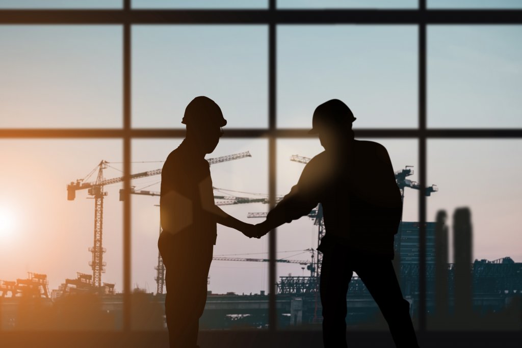 Silhouetted construction workers shake hands in front of large windows, with cranes and a sunset visible in the background.