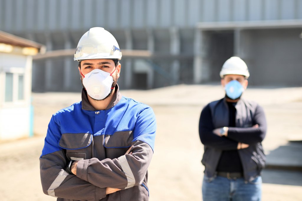 Two construction workers stand confidently outdoors, wearing safety helmets and serious expressions, against an industrial backdrop.