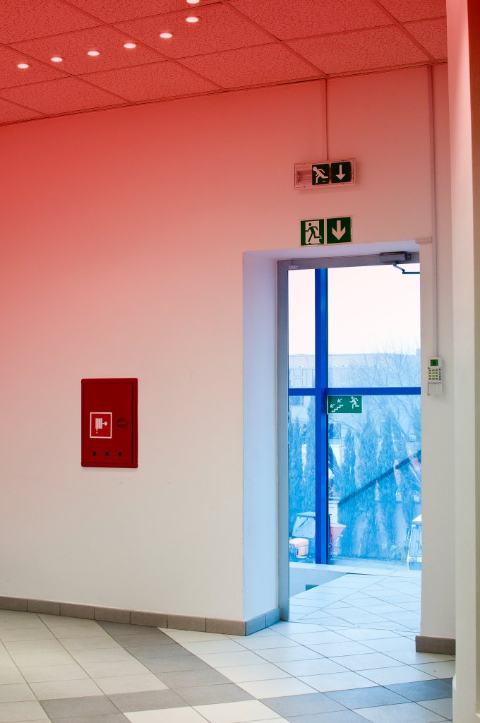 An exit door with blue glass, illuminated by soft red lighting, beside a red fire alarm panel and emergency exit signs.