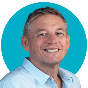 David Hornsby wearing a light blue shirt stands against a bright turquoise circular background, smiling gently with a neutral expression.
