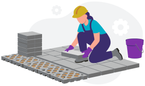 Worker in a hard hat and gloves kneeling to lay square paving stones on the ground with a stack of stones and a purple bucket nearby.